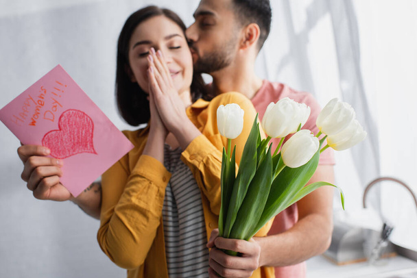 young man kissing girlfriend and presenting bouquet of flowers with greeting card in kitchen - Фото, изображение