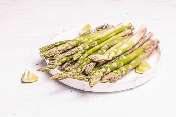 Ripe asparagus ready for cooking. Ceramic plate and lime pieces. Modern hard light, dark shadow. White wooden table, copy space - Foto, imagen