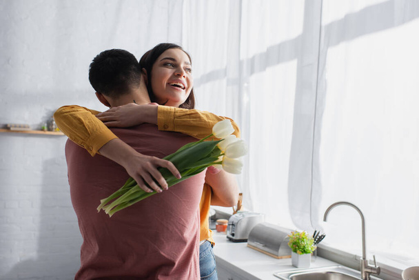 young man hugging smiling girlfriend with bouquet of flowers in kitchen - Photo, Image