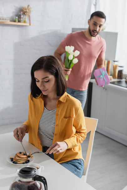 mujer joven desayunando cerca de novio borroso con ramo de flores y caja de regalo en la cocina - Foto, imagen