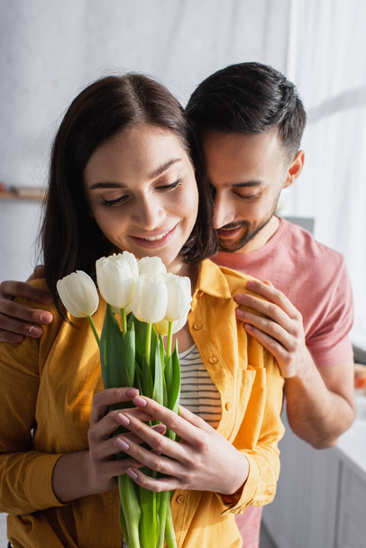 smiling young man hugging girlfriend with bouquet of flowers in kitchen - Фото, изображение