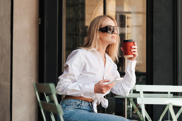 Elegancka kobieta siedzi przy stoliku kawiarni i trzymając filiżankę kawy, za pomocą smartfona, Pewna siebie kobieta przedsiębiorca w okularach przeciwsłonecznych i białej koszuli czeka na spotkanie na tarasie kawiarni. - Zdjęcie, obraz