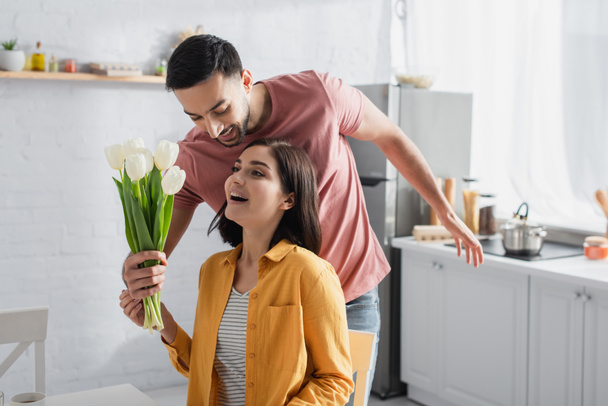 smiling young man presenting bouquet of flowers to excited girlfriend with open mouth in kitchen - 写真・画像