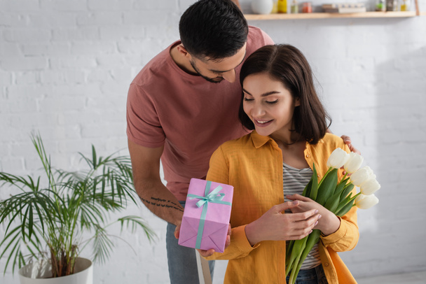 young man presenting gift box to girlfriend with bouquet of flowers in kitchen - Fotoğraf, Görsel