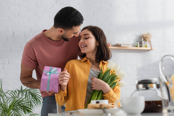 glimlachende jonge man presenteren geschenk doos aan vriendin met boeket van bloemen in de keuken - Foto, afbeelding