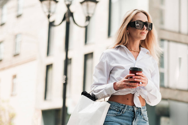 Elegant Blonde Woman Holding Cup of Coffee in Hand and Looking Away, Confident Female Entrepreneur Wearing Sunglasses and Golden Jewelry Walking on the Street of City. - Fotoğraf, Görsel