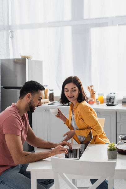 smiling young woman drinking coffee near boyfriend working with laptop in kitchen - Photo, Image