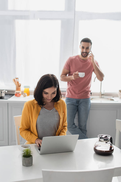smiling young woman working with laptop near blurred boyfriend speaking on cellphone in kitchen - Photo, Image