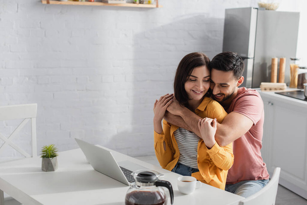 smiling young man hugging girlfriend with laptop in kitchen - Φωτογραφία, εικόνα