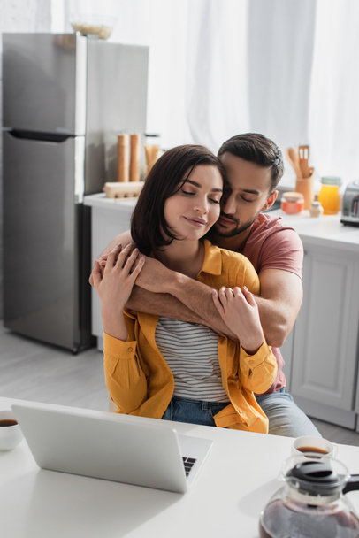 smiling young man with closed eyes hugging girlfriend with laptop in kitchen - Photo, Image