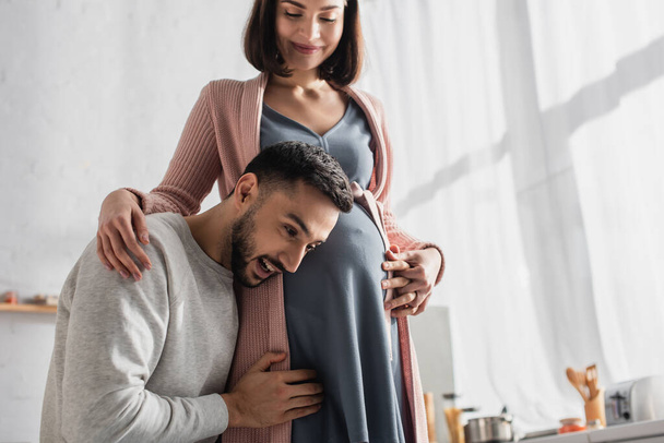 young man gently hugging pregnant woman and holding head near belly in kitchen - Photo, image