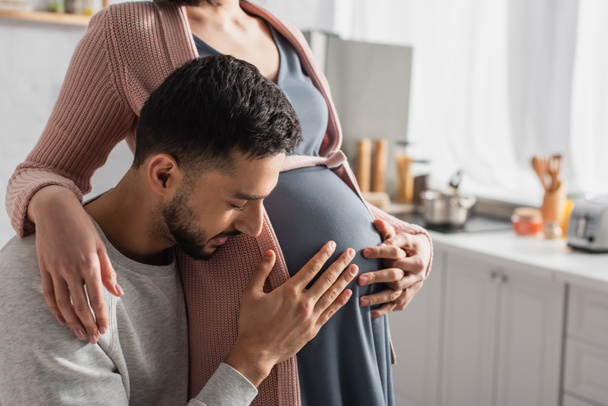 young man with closed eyes gently hugging belly of pregnant woman in kitchen - Photo, Image