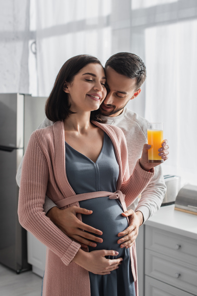 young man with closed eyes gently hugging pregnant woman with orange juice in kitchen - Фото, изображение