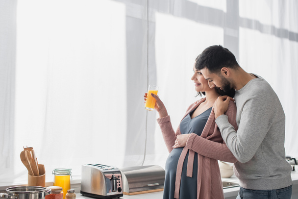 junger Mann mit geschlossenen Augen umarmt Schwangere in Küche sanft mit Orangensaft - Foto, Bild