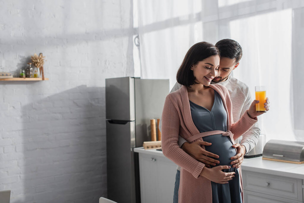 молодий чоловік з закритими очима ніжно обіймає вагітну жінку з апельсиновим соком на кухні
 - Фото, зображення