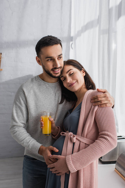 усміхнений молодий чоловік обіймає вагітну жінку апельсиновим соком на кухні
 - Фото, зображення