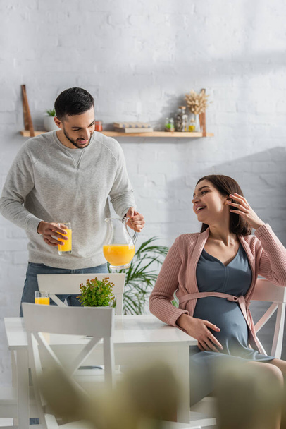 positive young man standing with jar and glass of orange juice near pregnant woman in kitchen - Photo, Image