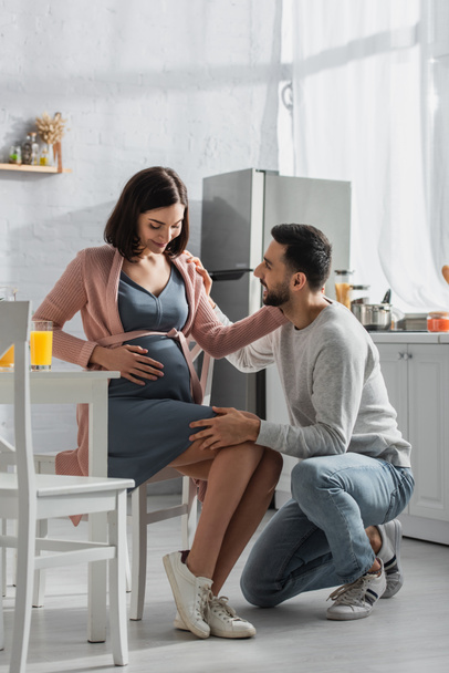 positive young man sitting on knee near pregnant woman in kitchen - Fotoğraf, Görsel