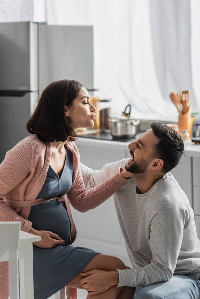 smiling young man sitting near pregnant woman with pouting lips in kitchen - Foto, Bild