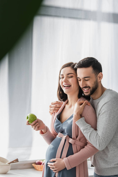 smiling young man with closed eyes hugging pregnant woman with apple in kitchen - Photo, Image