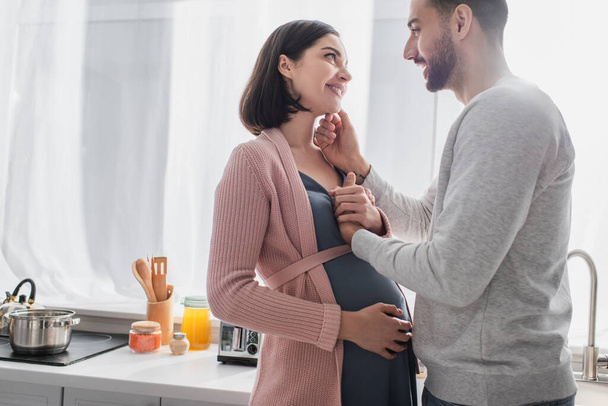 счастливый молодой человек трогает лицо беременной женщины на кухне - Фото, изображение