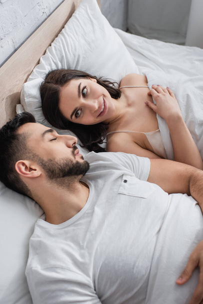 vista ad alto angolo di giovane donna sorridente che guarda il fidanzato addormentato a letto con biancheria bianca in camera da letto - Foto, immagini