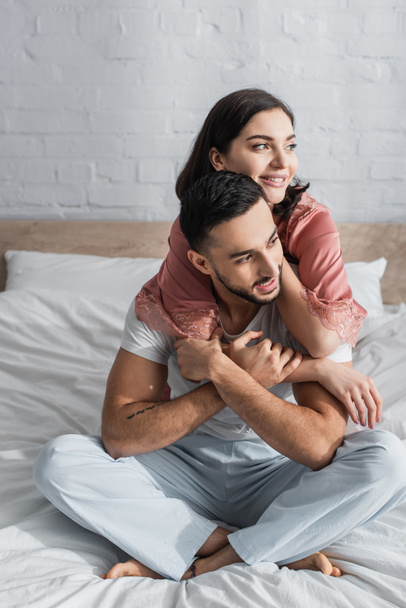 positive young couple sitting on bed with white linen and hugging in bedroom - Photo, Image