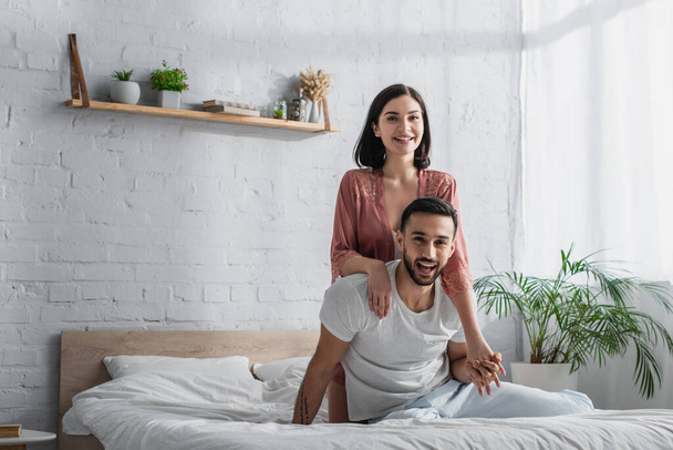 glückliches junges Paar sitzt mit weißer Bettwäsche auf dem Bett, blickt in die Kamera und umarmt sich im Schlafzimmer - Foto, Bild