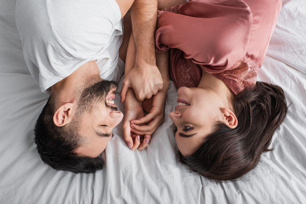 Blick von oben auf einen jungen Mann, der mit weißer Bettwäsche auf dem Bett liegt und seine Freundin im Schlafzimmer sanft an den Händen hält - Foto, Bild