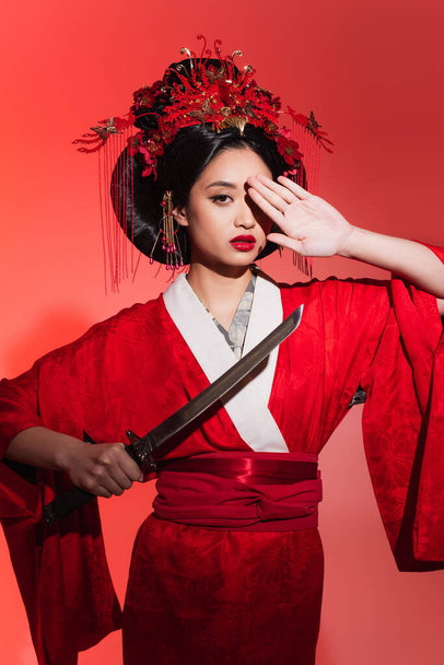 Asian woman in oriental clothes holding sword and covering eye on red background  - Foto, Imagem