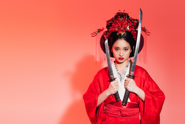 aziatische vrouw in authentieke kimono holding zwaarden op rode achtergrond  - Foto, afbeelding