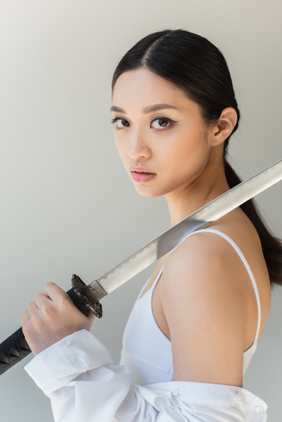 Asian woman in white clothes holding sword on shoulder isolated on grey - Foto, Imagen
