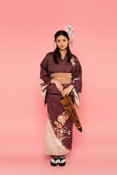 Japanese woman with traditional hairdo and kimono holding umbrella on pink background - Foto, afbeelding