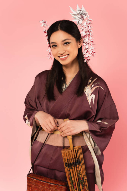 Gülümseyen Japon kadın pembe üzerinde el çantası ve şemsiye tutuyor.  - Fotoğraf, Görsel