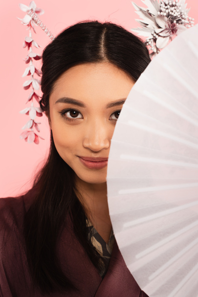 Mladá asijská žena při pohledu na kameru při držení ventilátor izolovaný na růžové  - Fotografie, Obrázek