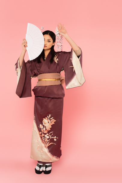 Full length of japanese woman in kimono holding fan near face on pink background - Фото, зображення