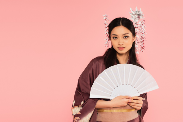 Bonita mujer asiática sosteniendo ventilador blanco y mirando a la cámara aislada en rosa  - Foto, Imagen