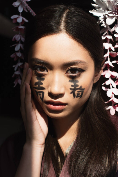 Young japanese woman with hairsticks and hieroglyphs on face isolated on black  - Photo, image