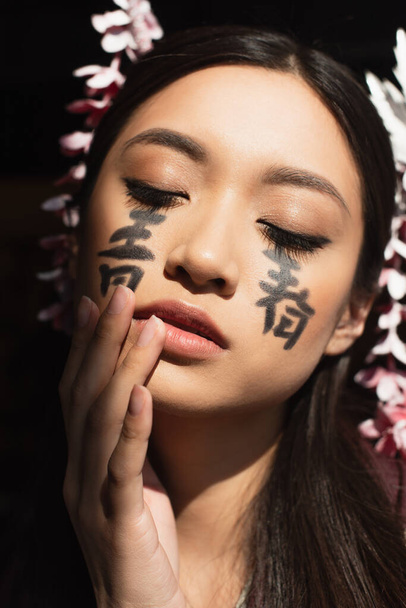Японская женщина с иероглифами на лице, стоящая с закрытыми глазами, изолированными на черном  - Фото, изображение
