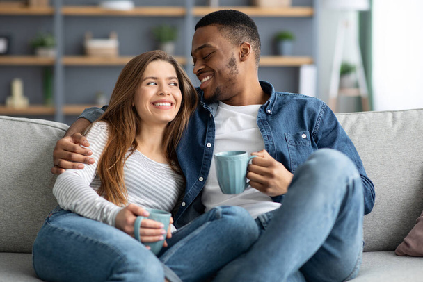 幸せな若い異人種間のカップルは自宅でコーヒーを飲み、カウチで抱擁 - 写真・画像
