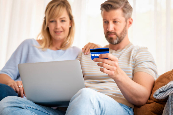 Online shopping from home. Handsome man holding bank card in his hand while her wife sitting next to him and using laptop while shopping online. Focus on bank card.  - 写真・画像