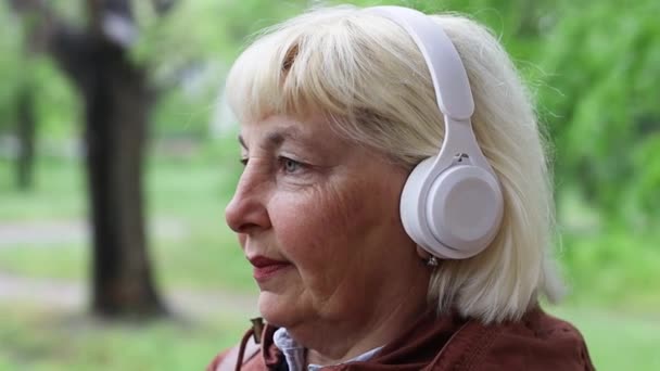 Vista de cerca de caucásico feliz 50 años de edad, mujer con auriculares blancos escuchando música con los ojos cerrados en la calle al aire libre. - Imágenes, Vídeo