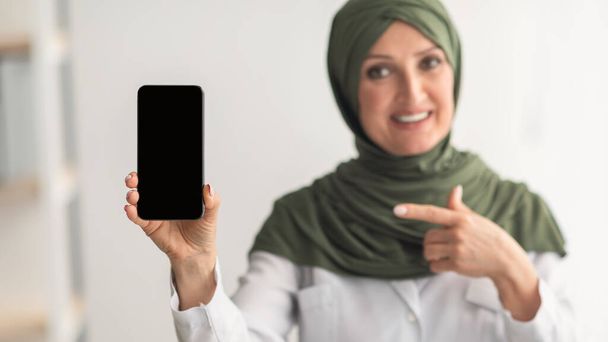 Мусульманская женщина-врач демонстрирует смартфон с чистым экраном в офисе - Фото, изображение