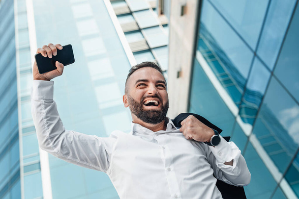 Mature job seeker celebrating successful interview raising fist with smartphone walking against glass business center - Фото, зображення