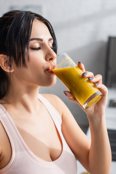 young woman holding glass and drinking smoothie in kitchen  - Foto, afbeelding