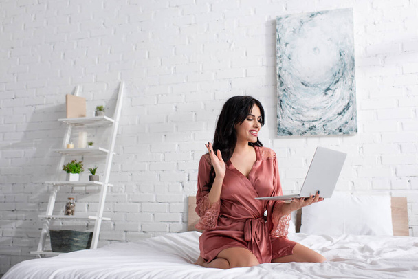 glückliche Frau im Seidenmantel winkt bei Videoanruf im Schlafzimmer  - Foto, Bild