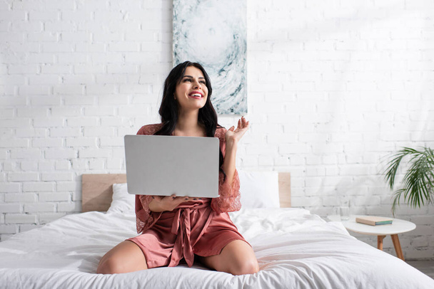 happy young woman in silk robe holding laptop in bedroom  - Фото, изображение