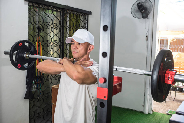 Um homem asiático apto em um boné branco se prepara para fazer agachamentos frente barbell em um ginásio em casa. Punho de braço cruzado. Exercício de perna e rotina de treinamento. - Foto, Imagem