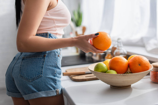 cropped view of young woman holding ripe orange in kitchen   - Foto, imagen