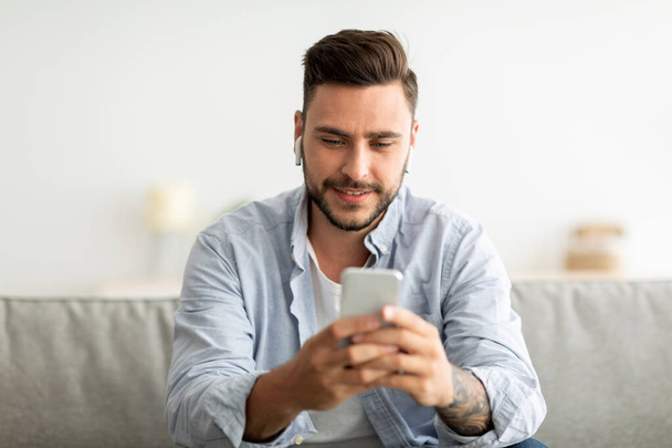 Young man with earbuds using smartphone, browsing internet and listening to music online, sitting on couch at home - Foto, Imagen